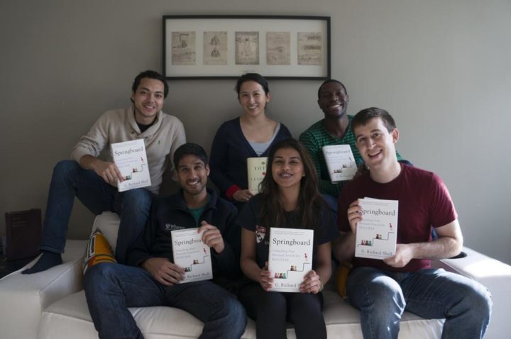 A group of people sitting on a couch, each holding a copy of the same book titled "Springboard." They are smiling and positioned in a casual setting with framed art on the wall behind them.