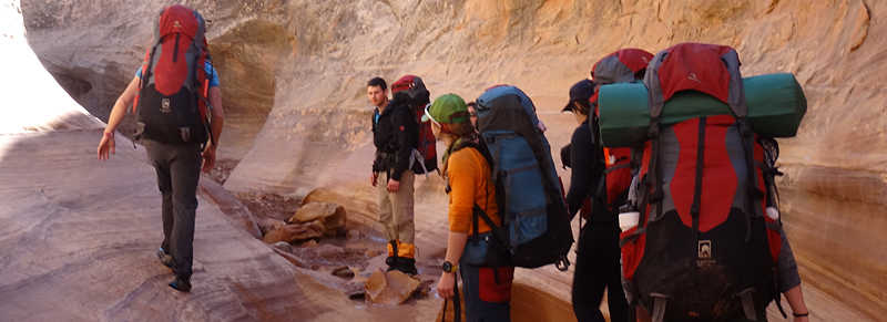 A group of people hiking in a rocky canyon, each carrying large backpacks with camping gear. The landscape features smooth, curved rock formations.