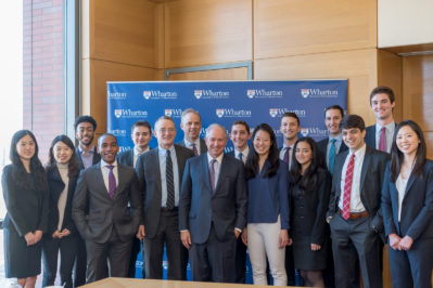 A group of people dressed in business attire standing in front of a backdrop displaying the Wharton School logo.