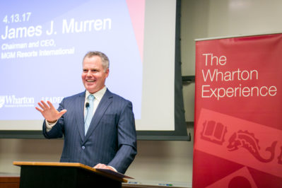 A person in a suit speaking at a podium, with a presentation screen and a banner reading "The Wharton Experience" in the background.