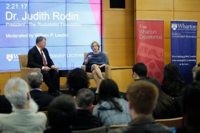 A speaker event with two individuals seated on stage in front of an audience. A large screen displays the text "Dr. Judith Rodin, President, The Rockefeller Foundation." Banners for The Wharton School are visible.