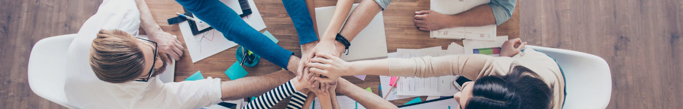 A group of people sitting around a table, placing their hands together in the center, symbolizing teamwork and collaboration. Papers and electronics are visible on the table.