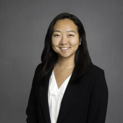 Headshot of a person with long black hair, wearing a white blouse and black blazer, smiling in front of a gray background.