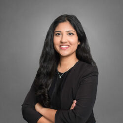 A person with long dark hair, smiling, arms crossed, wearing a dark blazer and a necklace, in front of a gray background. Headshot.