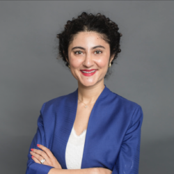 A person with curly hair wearing a blue blazer over a white top, smiling with arms crossed in front of a gray background.