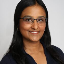 Headshot of a person with long dark hair and glasses, smiling, wearing a dark top against a plain background.