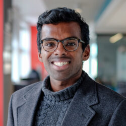 Headshot of a person wearing glasses and a dark sweater, smiling in an indoor setting with blurred background.