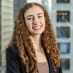 Headshot of a person with curly hair wearing a black blazer and smiling, set against an urban office background.