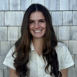 Headshot of a smiling person with long dark hair, wearing a light-colored button-up shirt, in front of a wooden shingle wall.