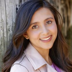 Headshot of a person with long, dark hair smiling in front of a weathered wooden surface. They are wearing a light-colored jacket.