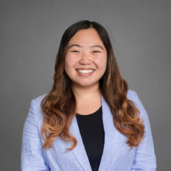 Headshot of a person smiling in a blue blazer and black top against a gray background.