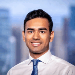Headshot of a person smiling, wearing a white shirt and blue tie, with a blurred cityscape in the background.