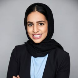 Headshot of a smiling person wearing a black hijab and a dark blazer over a light blue top. They are posed against a plain background.
