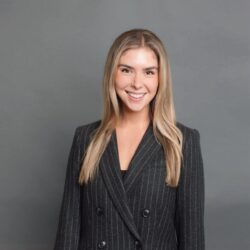 A person in a professional headshot wearing a dark pinstripe suit, smiling in front of a gray background.