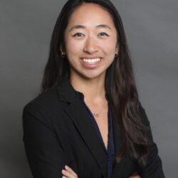 A professional headshot of a person with long dark hair, smiling, wearing a black blazer and dark top, set against a plain background.