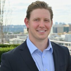 A person in a suit and light blue shirt smiling, with a cityscape and sky in the background.
