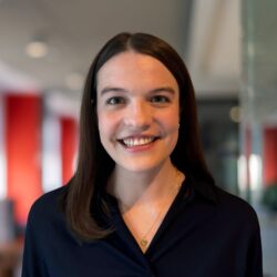 Headshot of a person with long, straight hair, smiling indoors. The background is blurred with red and white elements.