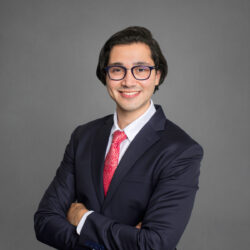 Headshot of a person wearing glasses, a dark suit, and a red tie, with their arms crossed, set against a plain background.