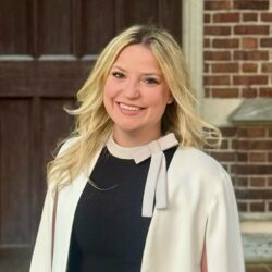 Headshot of a smiling person with long blonde hair, wearing a black top and a white jacket. A brick wall and wooden door are in the background.
