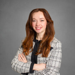 Headshot of a person with long auburn hair, wearing a plaid jacket and black top, posing with arms crossed against a gray background.