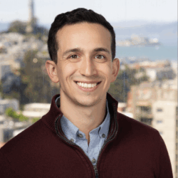 Headshot of a smiling person with short dark hair, wearing a maroon sweater and a blue shirt. The background shows an out-of-focus cityscape with water and buildings.