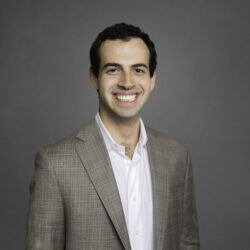A person in a plaid suit jacket and white shirt smiling against a gray background; a professional headshot.