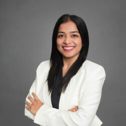 Headshot of a person with long black hair, smiling, wearing a white blazer and black top against a gray background.