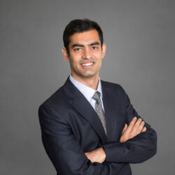 A professional headshot of a person in a dark suit with a light-colored shirt and polka dot tie, arms crossed, smiling against a gray background.