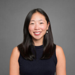 Headshot of a person smiling, with long, straight hair, wearing a sleeveless dark top and pearl earrings, set against a plain background.