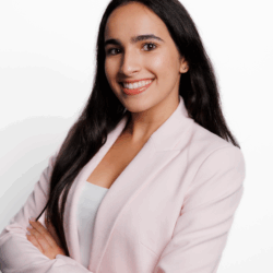 Headshot of a smiling person with long, dark hair wearing a light pink blazer and white top, posed against a white background.
