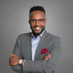 A person in a gray suit and glasses, smiling confidently with arms crossed, wearing a watch and red pocket square, set against a plain background.