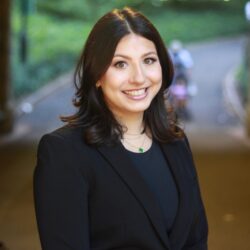 Headshot of a person with dark hair wearing a black blazer, smiling, standing outdoors with greenery in the blurred background.