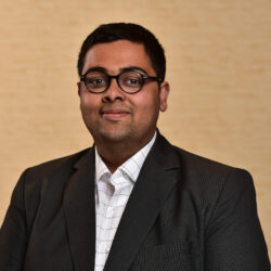 Headshot of a person wearing glasses, a dark suit jacket, and a white checkered shirt, standing against a plain background.