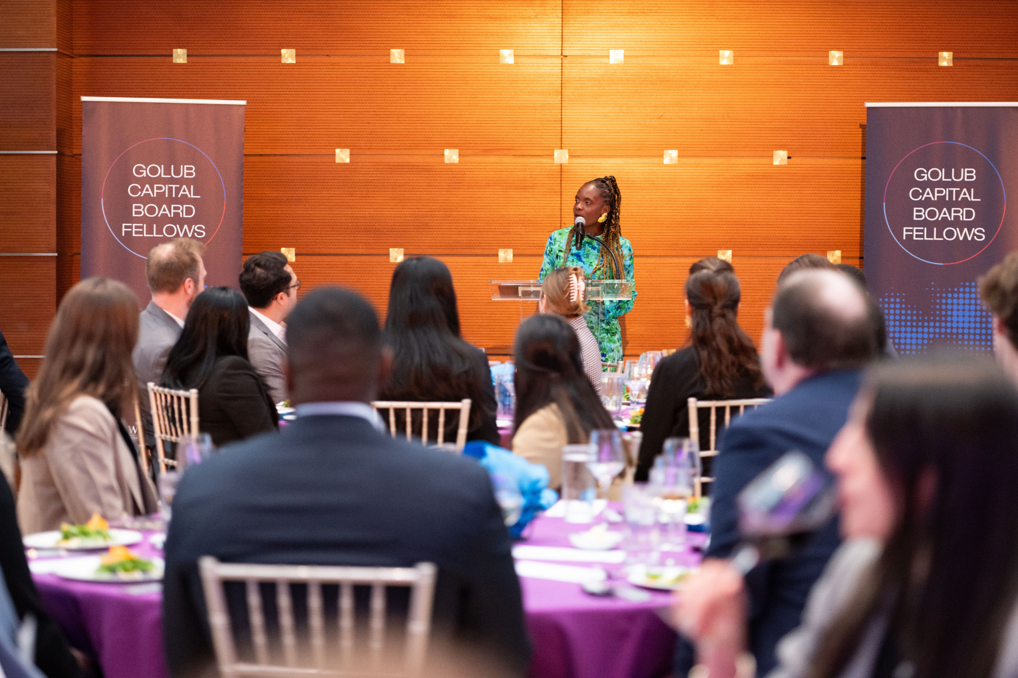 A speaker stands at a podium addressing an audience at an event with "Golub Capital Board Fellows" banners visible. Several people are seated at round tables with purple tablecloths.