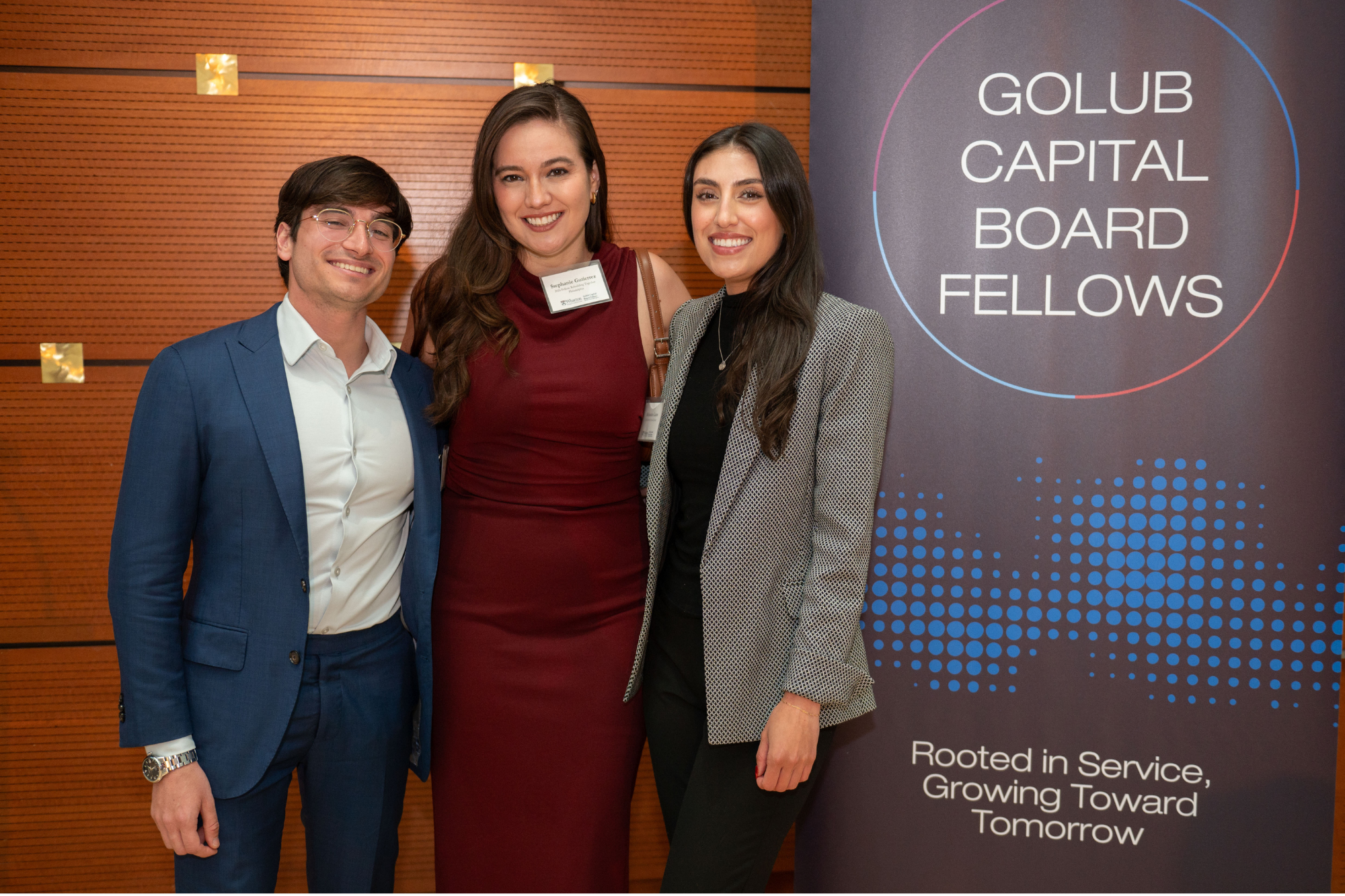 A group portrait of three people, two in business attire and one in a formal dress, standing beside a banner that reads "Golub Capital Board Fellows."