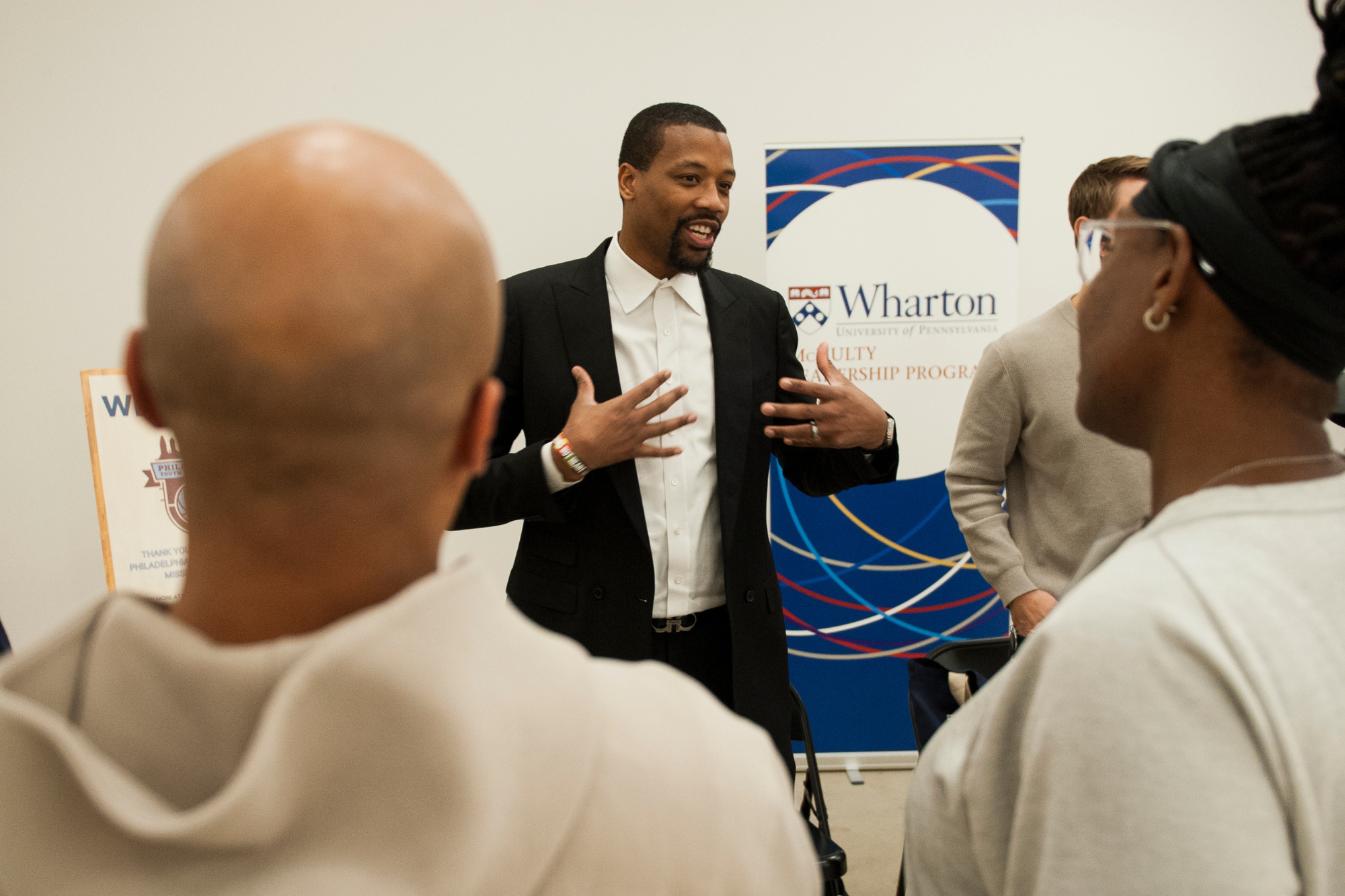 A person is speaking to a small group of people in front of a Wharton School banner.