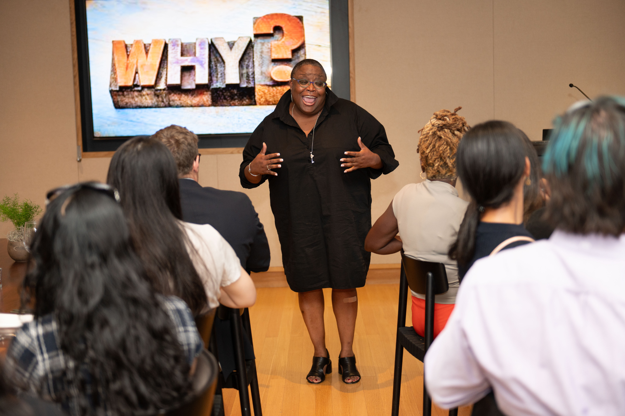 A speaker is presenting to a small group of people in a room. A screen behind the speaker displays the word "WHY?".