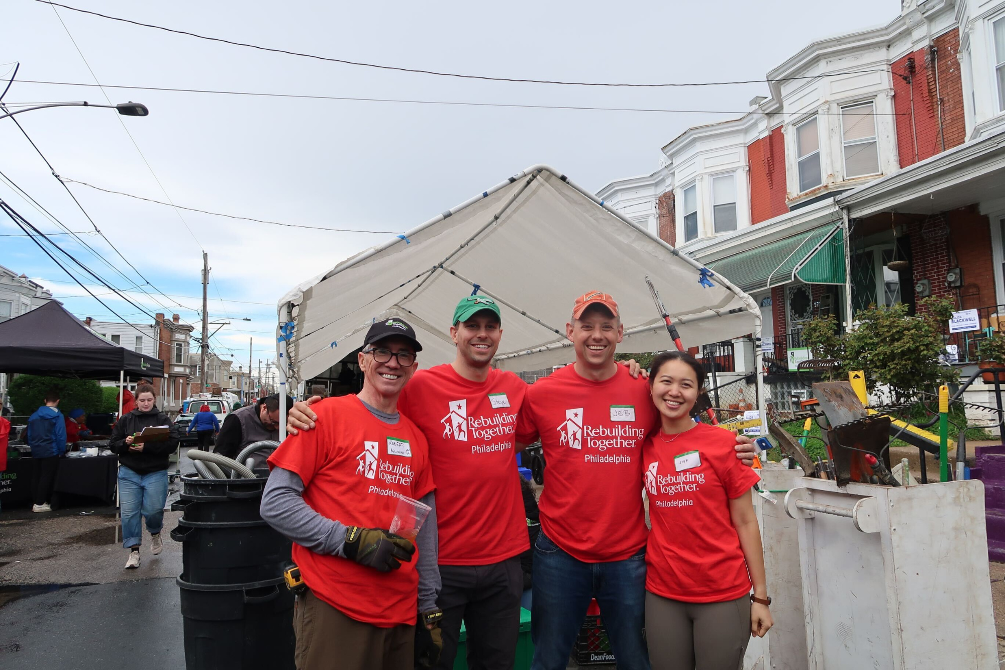 A small group of people in matching red "Rebuilding Together Philadelphia" t-shirts stand smiling under a canopy on a residential street. They appear to be participating in a community or volunteer event.