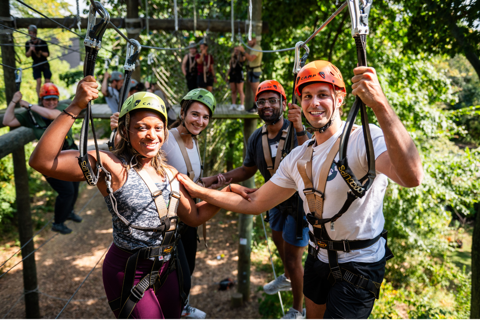A group of people wearing helmets and harnesses engaging in a zip-lining or ropes course activity surrounded by trees. They appear happy and excited.