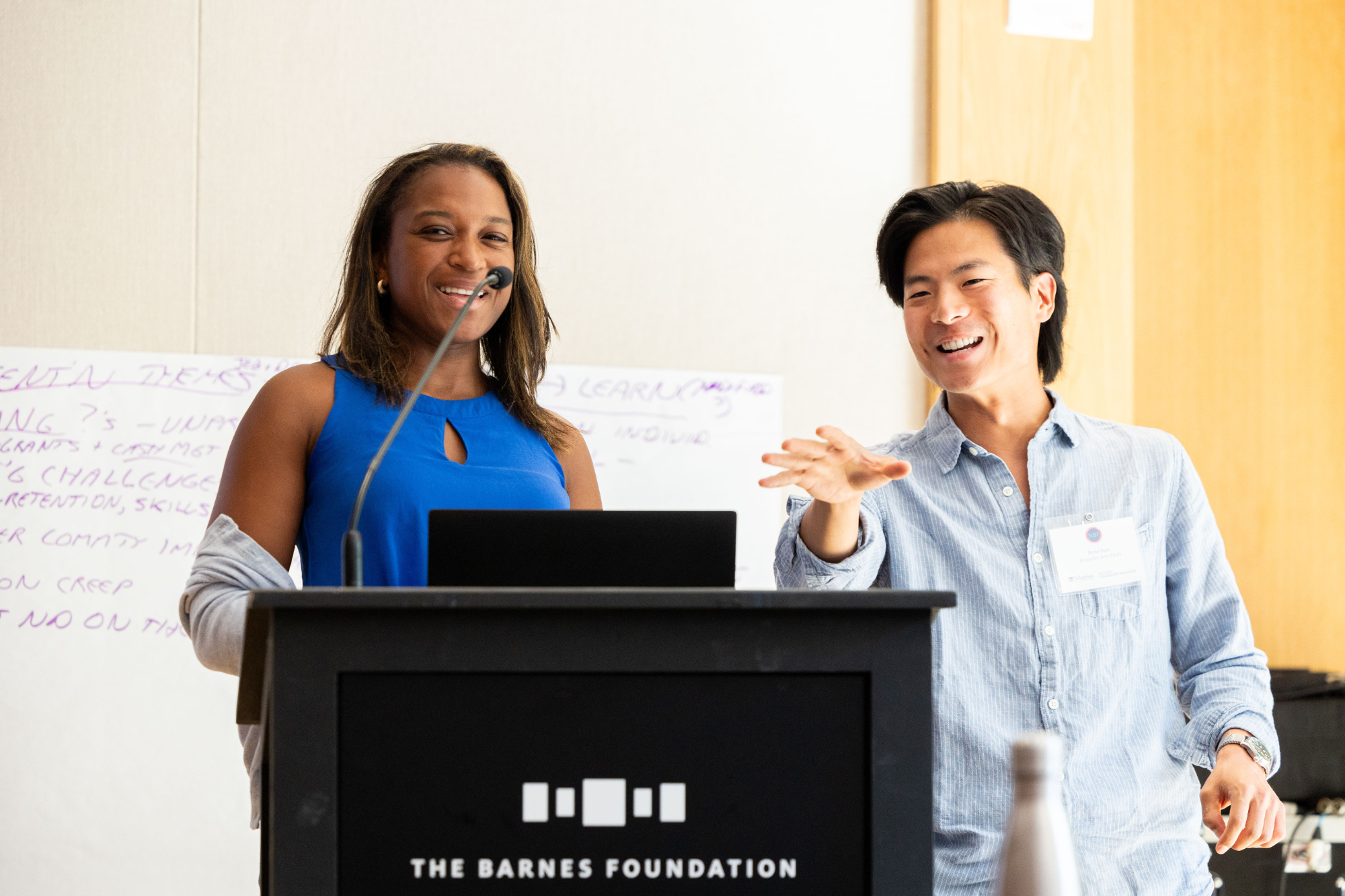 A person wearing a blue sleeveless top stands at a podium with "The Barnes Foundation" logo, smiling into a microphone, while another in a light shirt gestures beside them.