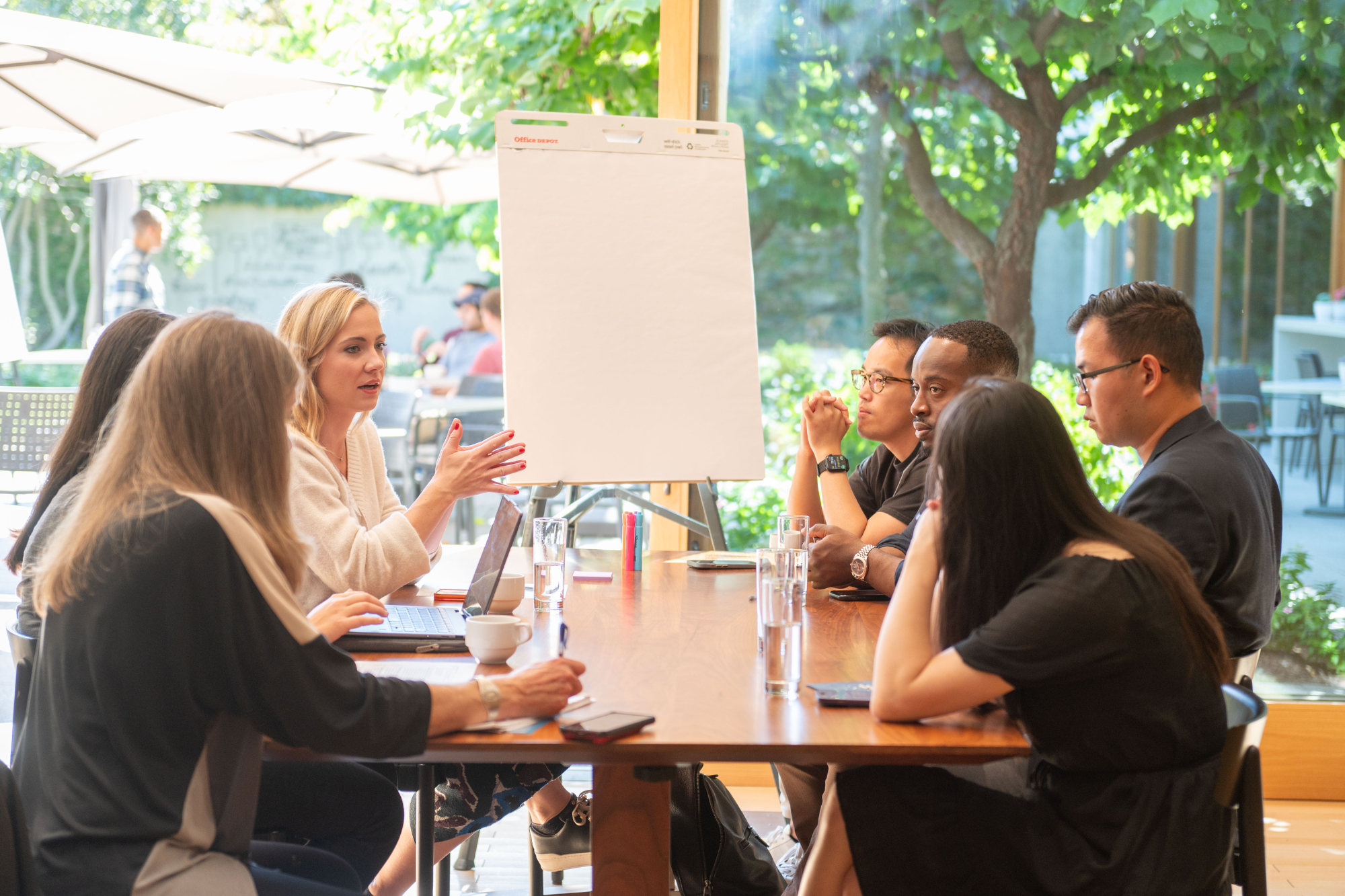 A small group of people engaged in a meeting around a wooden table. One individual is speaking with gestures, while a flip chart stands in the background. The setting appears to be a bright, outdoor or garden-like space with natural light.