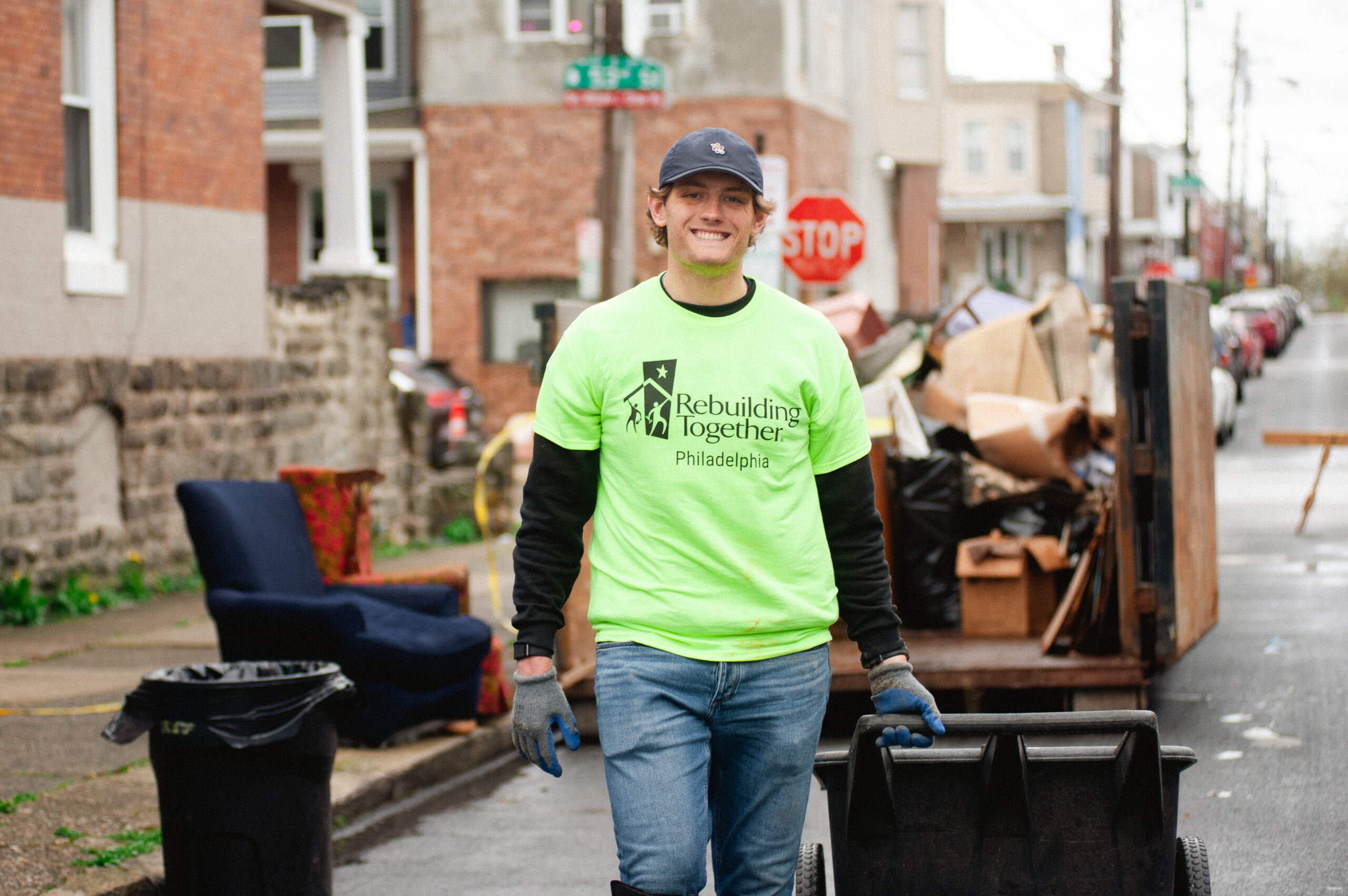 A person wearing a bright green "Rebuilding Together Philadelphia" shirt and gloves smiles while pushing a trash bin on a street. There is a pile of debris in the background.