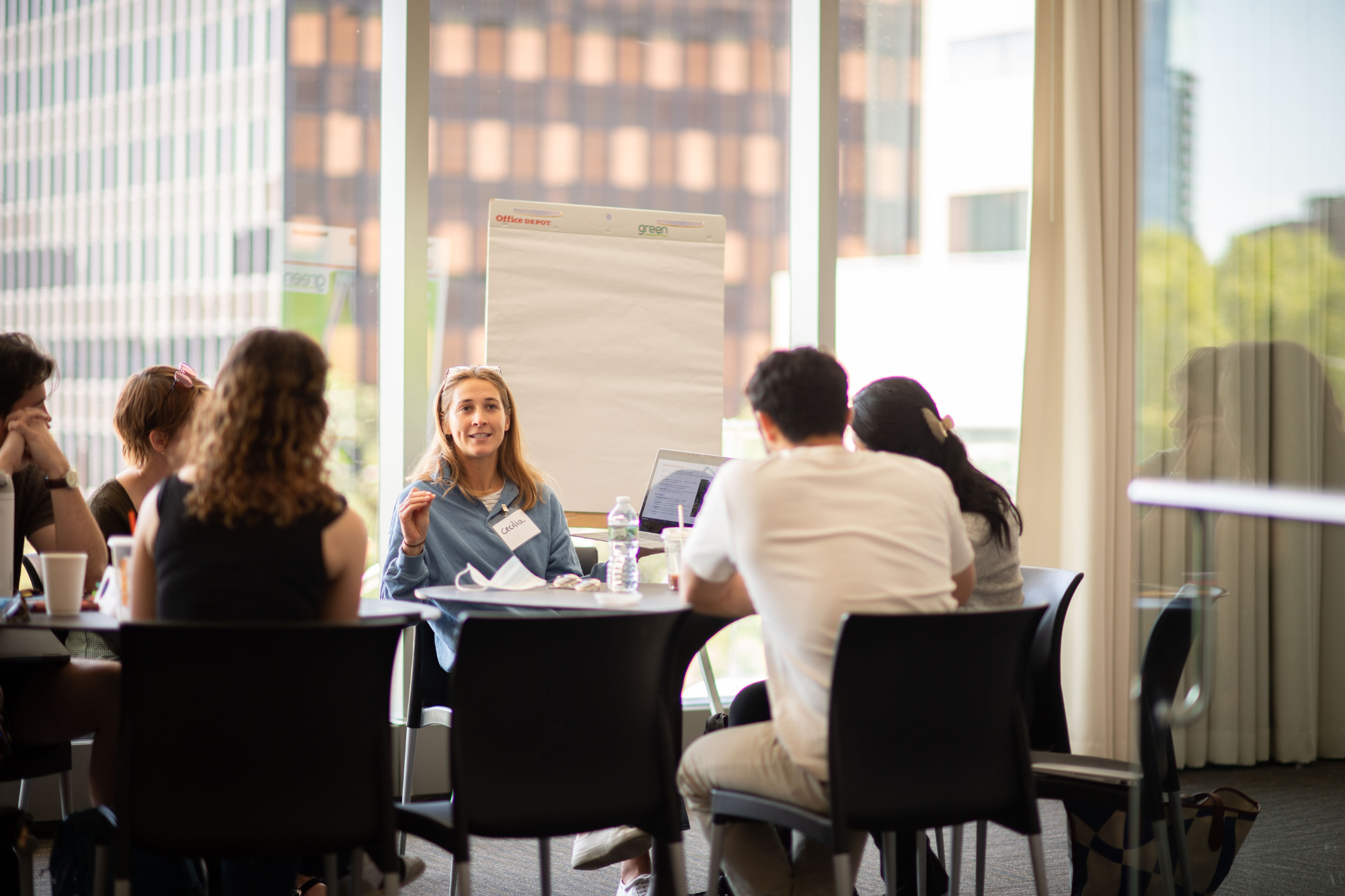 A small group of people engaged in a discussion around a table, with a person speaking and a notepad on a stand in the background. They appear to be in a modern meeting or conference room with large windows.