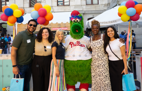 A group of people smiling and posing with the Philadelphia Phillies mascot in a lively outdoor setting. Colorful balloons and festive decorations are visible in the background.