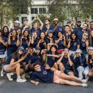 A large group of people wearing matching blue shirts and tennis attire, posing energetically in front of a building with a banner referencing leadership and education.